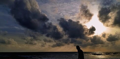A silhouette of a person wearing a cap stands at the edge of the ocean. Above, the sky is filled with large, dramatic dark clouds. Sunlight breaks through on the right, casting golden rays and illumin © Eka