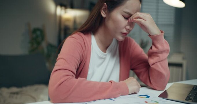 Exhausted Asian businesswoman pinching bridge of nose with eyes closed, suffering from eye strain at home.