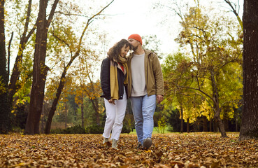 Naklejka premium Couple walking in autumn park among leaves. He kisses her forehead, sharing happy warmth as they stroll together through golden foliage and nature. Love, romance, and peaceful autumn together.