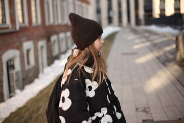 Cute young girl in a stylish black and white floral puffer jacket and brown beanie hat walking on a city street, profile view. © Irina