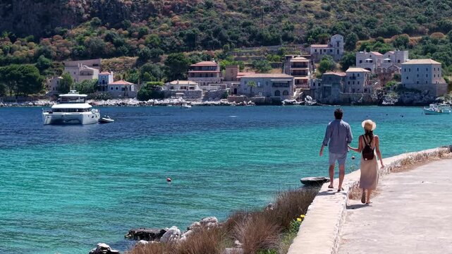 Couple walking hand in hand by the turquoise seashore promenade, Yachting in Limeni village, traditional stone buildings and mountains in the background, Static shot