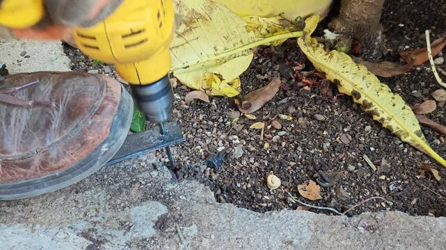 Yellow power drill bit piercing a steel flat bar held down on the floor by a foot