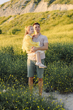 Father holding daughter in yellow dress in a sunny meadow near a coastal landscape.