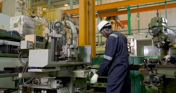 African adult male technician operates CNC grinding machine as coworker enters nearby, supporting production setup, machine monitoring and coordinated factory workflow in slow motion