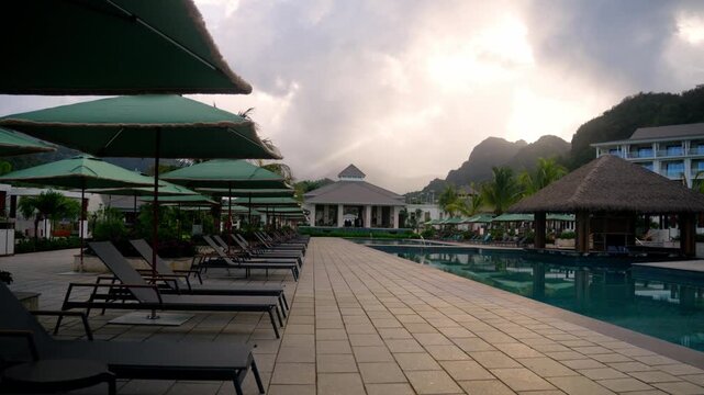 Camera pan across a luxury resort pool deck at twilight. The shot features rows of sun loungers under green umbrellas, a serene swimming pool, and the resort's main building set against a mountainous