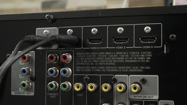 Hand connects an HDMI cable to one of several HDMI inputs on a home theater AV unit, revealing detailed labels and legacy video jacks.