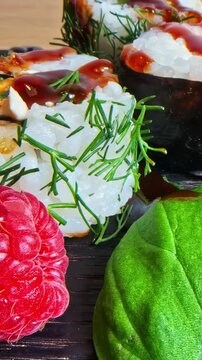 Bamboo chopsticks pick a sushi roll topped with salmon roe, fresh dill and teriyaki sauce from a dark wooden board surrounded by raspberries and a large green leaf in close up static shot