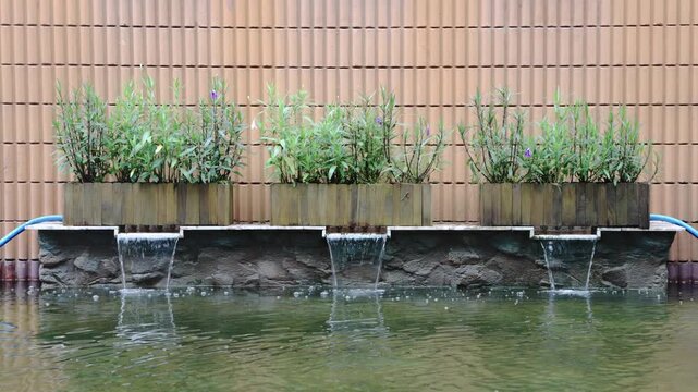 three wooden planter boxes over a stone base. Water cascades into a clear pond. Features green plants with small purple flowers against a tan textured wall.