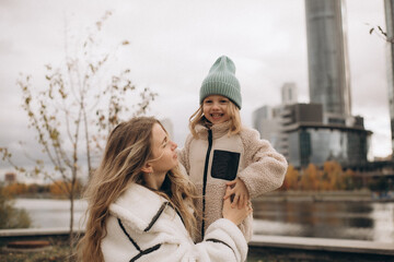 Happy young mother holding her laughing little daughter in a fleece jacket and beanie hat outdoors on a cloudy autumn day. © Irina