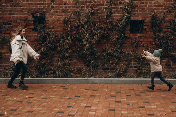 Happy young mother and little daughter in a beanie hat running towards each other for a hug against a red brick wall in autumn. © Irina