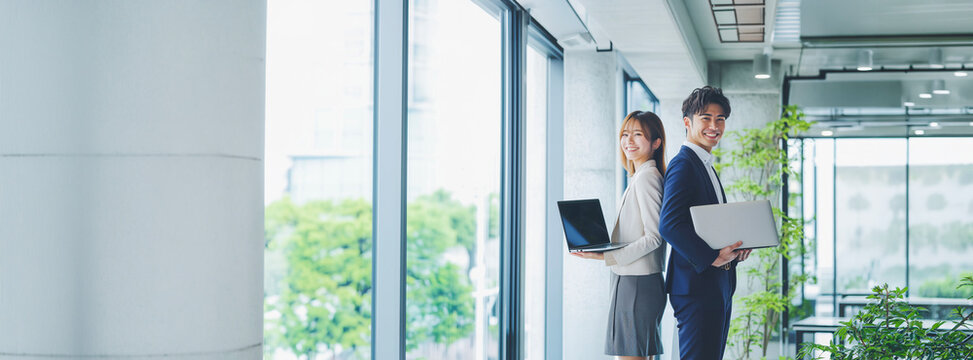 A smiling young businessman and businesswoman standing back-to-back holding laptops near a window with fresh greenery in a modern office.