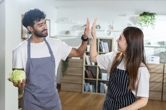 Happy couple cooking in home kitchen wearing apron sharing joyful high five gesture holding fresh green cabbage preparing vegetarian meal together celebrating successful teamwork with food