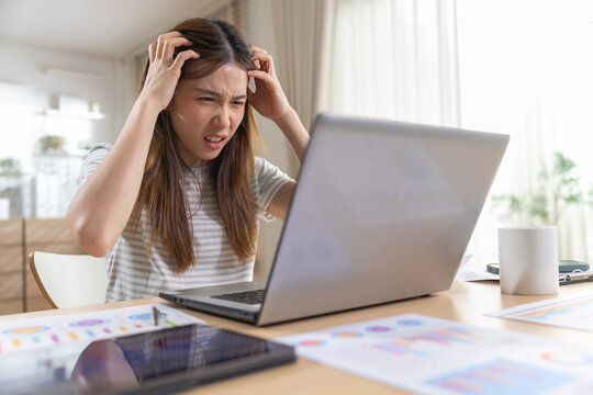 Stressed Asian woman working on laptop at desk feeling frustrated and angry pulling her hair over difficult project with business charts in home office