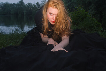 Mysterious red haired woman in a flowing black dress at the edge of a quiet lake at dusk. Dramatic portrait with an intense gaze, cinematic atmosphere and dark fairytale mood © ullision