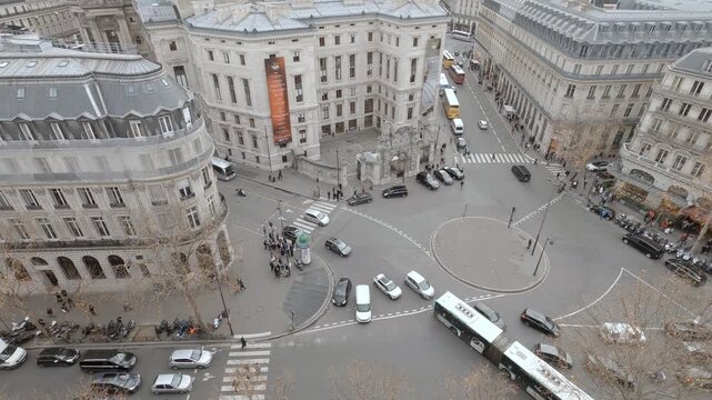 Elevated moving shot of the Palais Garnier and nearby Paris streets, with buses, cars and pedestrians circulating around the historic opera house on an overcast day.
