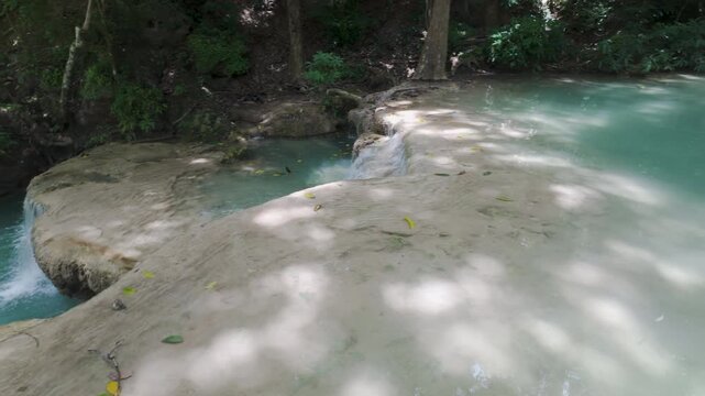 Pale mineral ledges border a blue-green stream as gentle water spills over the edge beneath dappled jungle shade.