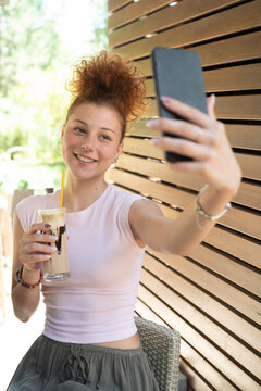 A young woman taking a selfie with a mobile phone, drinking coffee in a cafe, and smiling