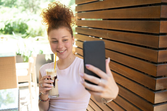 A young woman taking a selfie with a mobile phone, drinking coffee in a cafe, and smiling