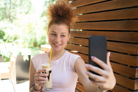 A young woman taking a selfie with a mobile phone, drinking coffee in a cafe, and smiling