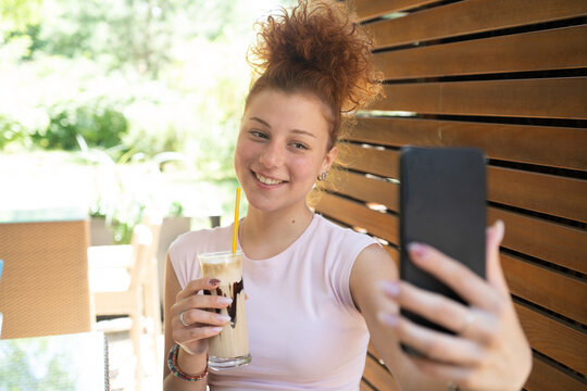 A young woman taking a selfie with a mobile phone, drinking coffee in a cafe, and smiling