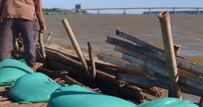 close up of workers lay halved bamboo stalks along the edge of a bamboo bridge under construction, stabilized by sandbags, with a wide river and a distant concrete bridge in the background.