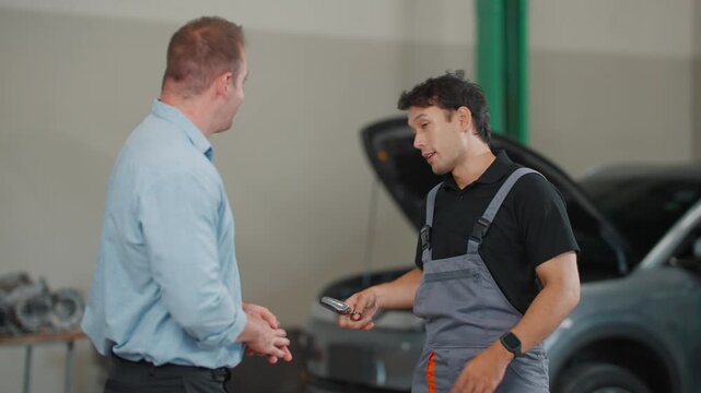 Two men converse near an open car hood. One wears overalls, gesturing as he explains. The other listens, back turned toward the camera. Professional interaction suggests a mechanic discussing repairs