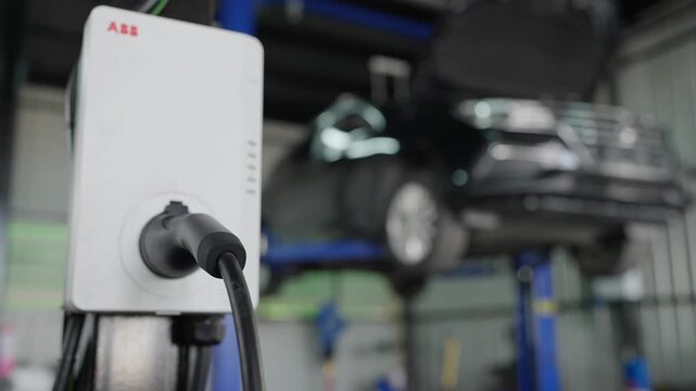 White charger stands in EV garage bay. Black cable plugs into charging port. Car hangs on lift in background. Toolroom lights shine overhead. Mechanic tools rest nearby