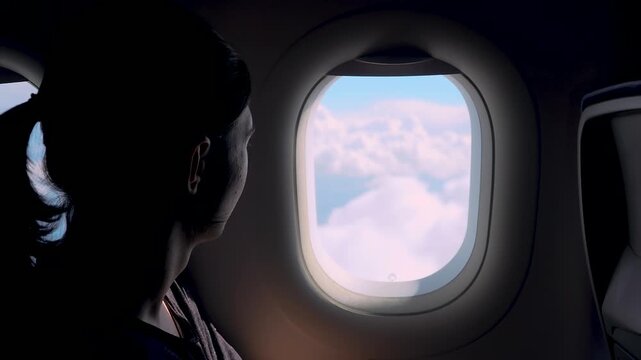 Passenger looking through airplane window at clouds during descent before landing. Young man watching soft cloud layers from aircraft seat in dim cabin. Aircraft gradually descending toward