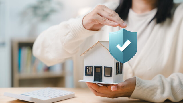 Home insurance and property protection concept shown with female hands holding model house near blue shield icon representing secure housing coverage and financial risk management