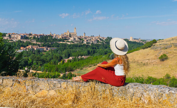 Rear view of young woman tourist in straw hat and red pants sitting on stone wall looking at panoramic skyline of Segovia, Spain