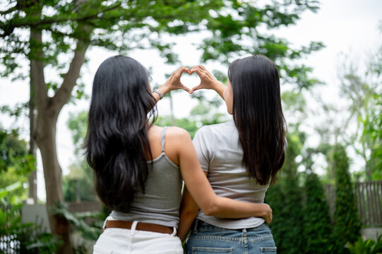 Rear view of two Asian women hugging each other and making a heart shape hand gesture in the park.