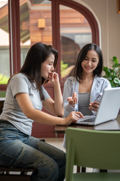Asian women coworkers collaborating with laptop while reviewing business information in modern cafe.