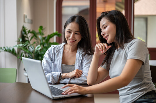 Smiling Asian woman typing on laptop while working with her teammate in cafe.