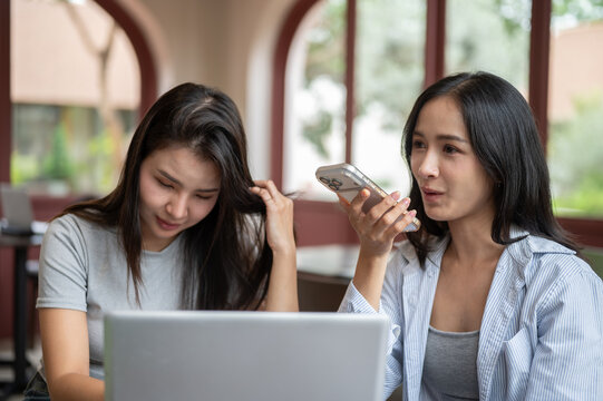 Asian woman talking or using voice assistant on smartphone beside friend while while working in cafe