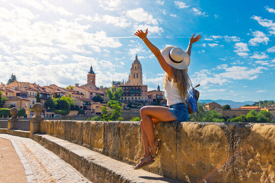 Happy young woman tourist with arms raised celebrating on stone wall with panoramic view of Segovia skyline in Spain