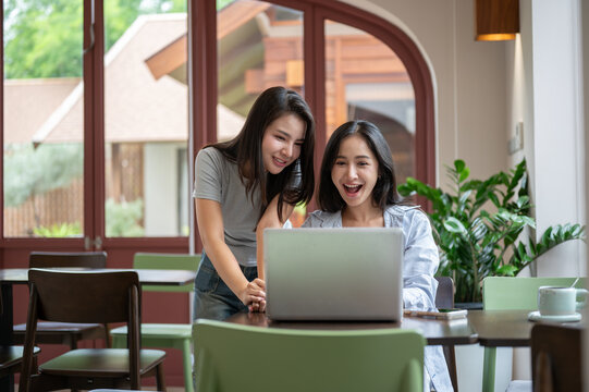 Asian women coworkers smiling at laptop after completing successful teamwork in a bright modern cafe