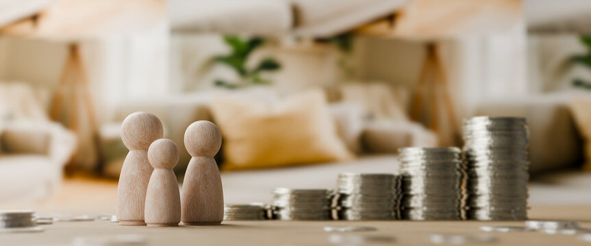 Wooden family figurines standing beside a growing stack of silver coins on a table with a soft focus home blurred interior background for financial growth