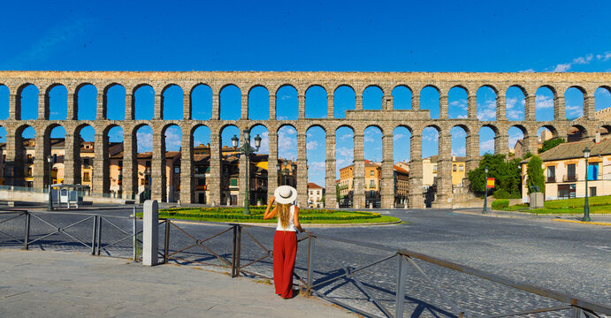 Rear view of young woman tourist in straw hat and red pants looking at ancient Roman aqueduct in Segovia, Tourism in Spain