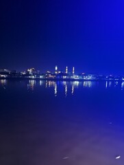 Fototapeta premium Nighttime view of a city skyline with illuminated buildings and their reflections shimmering on the calm water surface under a dark blue sky.