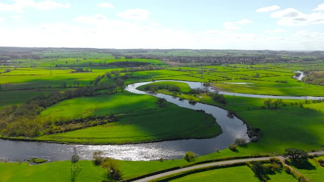 Sunlit river bend cutting across patchwork fields with strong light contrast over grassy terrain. Drone perspective farmland patterns water reflections and rolling countryside stretching to horizon