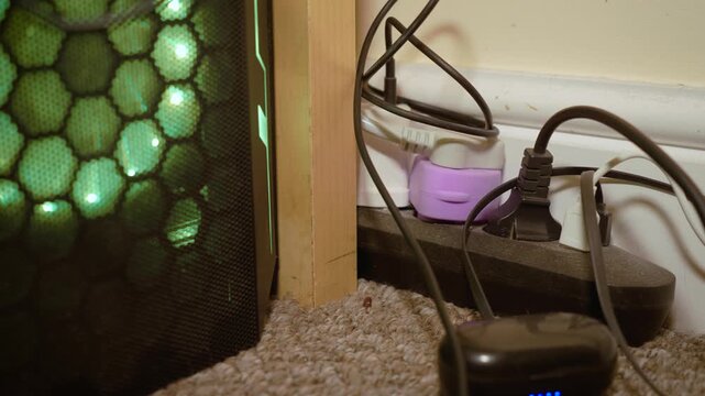 Gaming computer next to messy power strip with tangled charging cables. Floor level view revealing disorganized wiring and crowded electrical outlets. Home office cable management, technology clutter