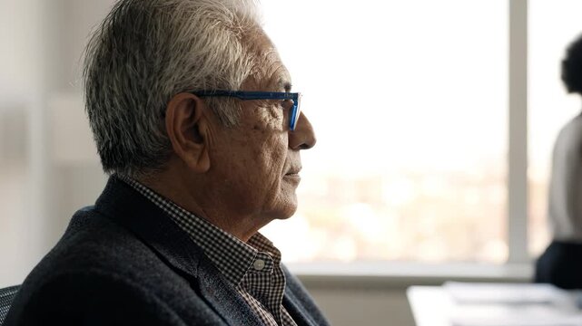 Elderly man in profile wearing glasses in modern office, African American woman with rainbow headband walking in background