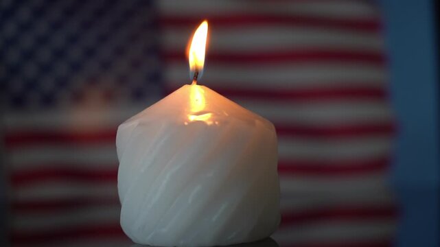 Single white candle burns brightly. Its flame glows against blurred American flag stripes. The candle's twisted shape reflects warm light. This moment evokes patriotism and remembrance