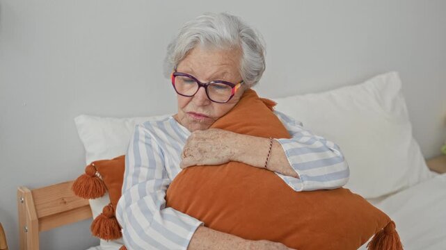 Woman hugging pillow on bed inside building, holding orange cushion and showing hands and glasses; quiet loneliness.