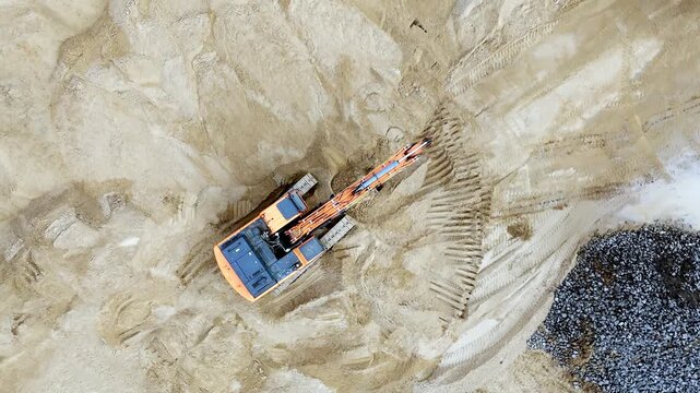 Aerial shot captures excavator digging sandy terrain. Orange and blue machine moves dirt into a deep hole. Tire tracks form patterns across the loose soil