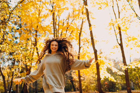 Smiling young woman plays and jumping in the autumn park with the yellow leaves at sunset.