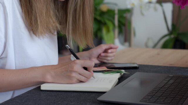 Woman planning home budget with phone writing notes in a notebook with euro cash and laptop on the table. Concept of household expenses and bills.