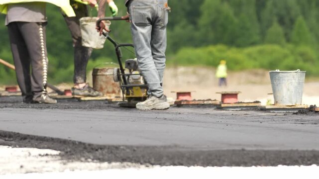 Workers wear yellow vests. One carries a bucket. Another pushes a compactor. Fresh asphalt covers the road. Green trees form the background. This footage suits road construction projects