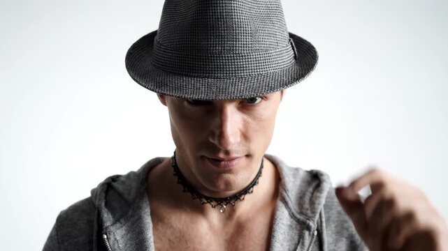 Stylish young man in gray fedora and hoodie tipping hat with confident gaze against white studio background