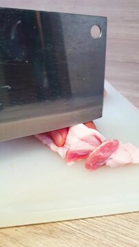 A person using a large kitchen cleaver knife to slice raw pork belly on a white cutting board for cooking